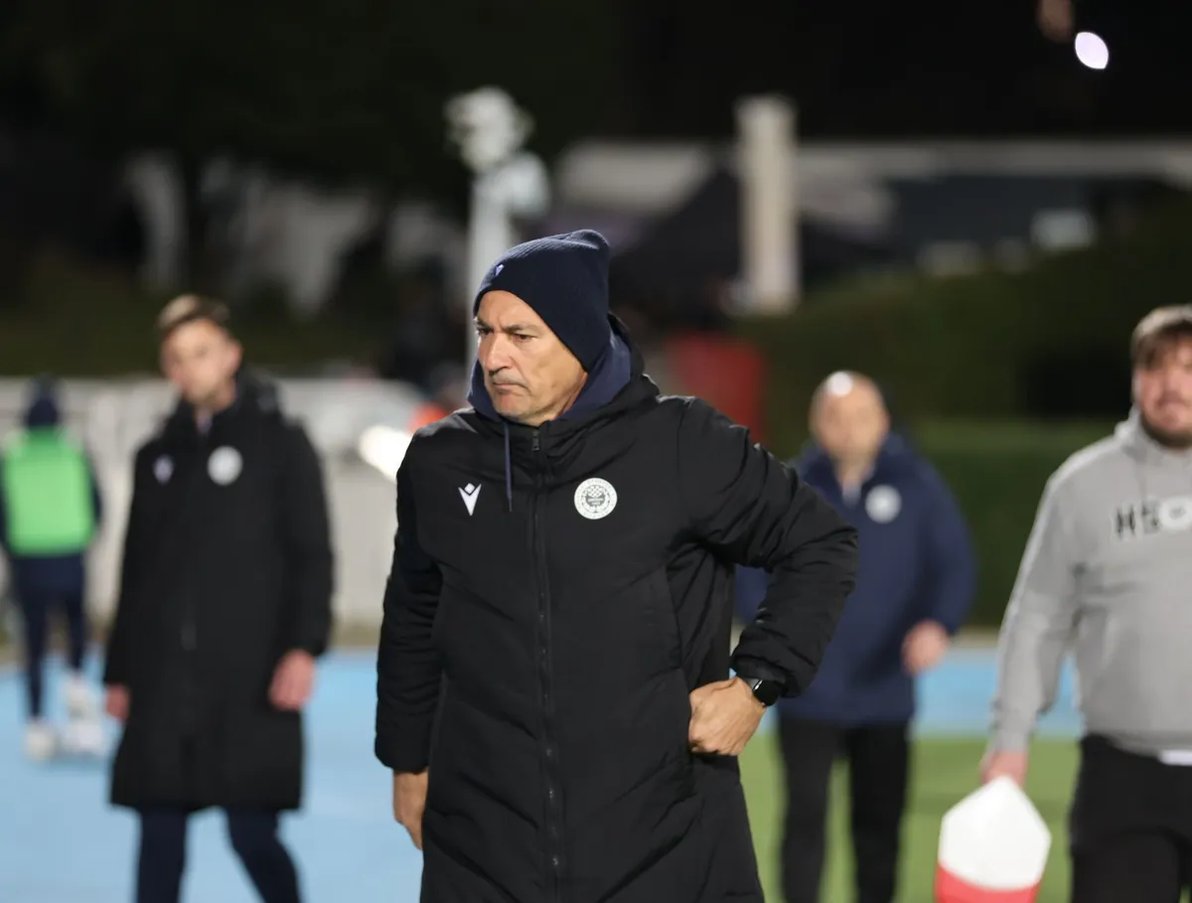 Fudbalski trener u crnoj jakni i plavoj kapi gleda u daljinu.