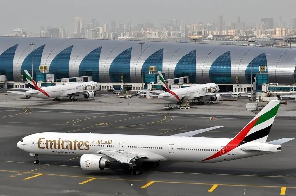 Avion Emirates na pisti aerodroma sa drugim avionima u pozadini i modernom zgradom terminala.