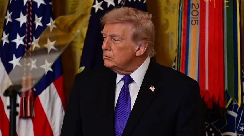 Donald Trump u crnom odijelu sa plavom kravatom, stoji ispred američke zastave i drugih zastava.