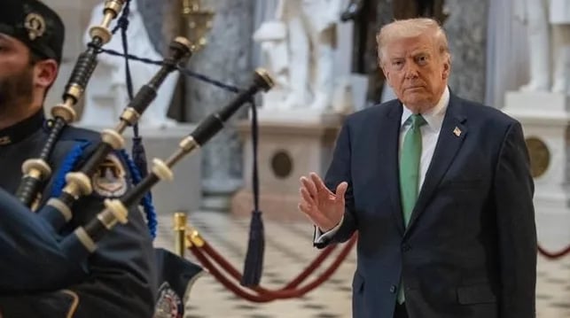 Donald Trump nosi tamno odijelo sa zelenom kravatom dok mu svira gajde u pozadini.