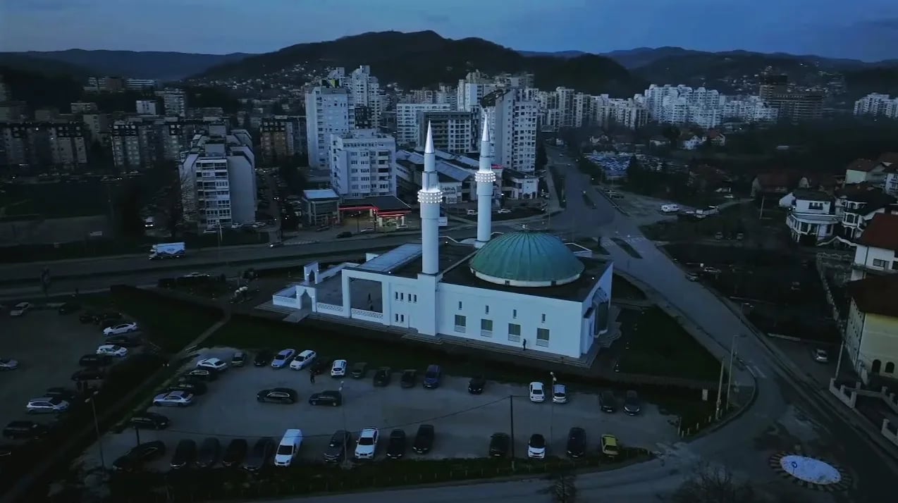 Moderna džamija sa zelenom kupolom i dva minareta okružena stambenim zgradama i planinama u sumrak.