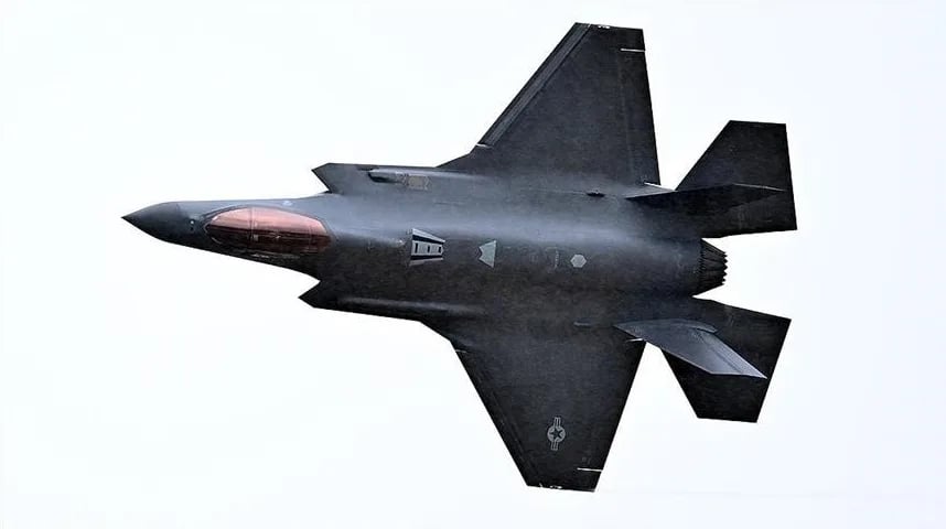 Crni F-35 borbeni avion sa pilotinom u boji ćilibara leti uvis.