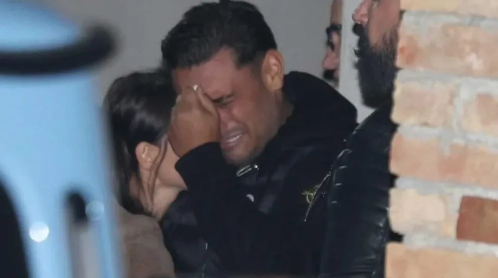 Čovjek sa rukom na čelu i suzama u očima, zagrljen od strane žene, dok ga sa desne strane dodiruje drugi muškarac sa bradom.