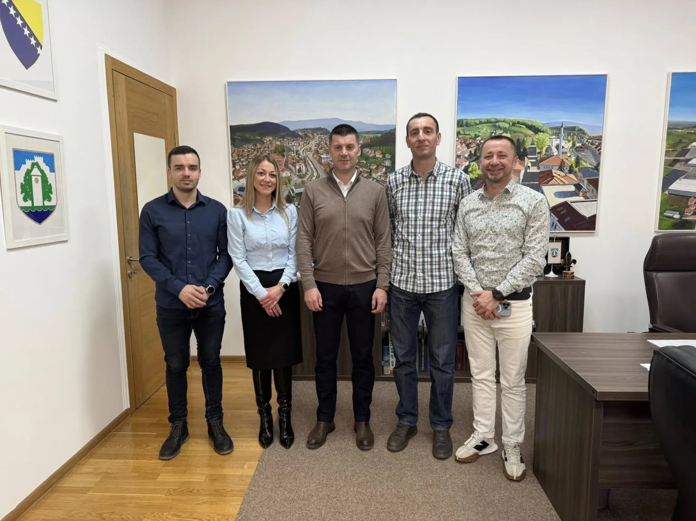Pet osoba, tri muškarca i dvije žene, pozira u moderno uređenoj kancelariji. Na zidovima se nalaze umjetničke slike gradova i grbovi. Osobe su odjevene u poslovnu odjeću.