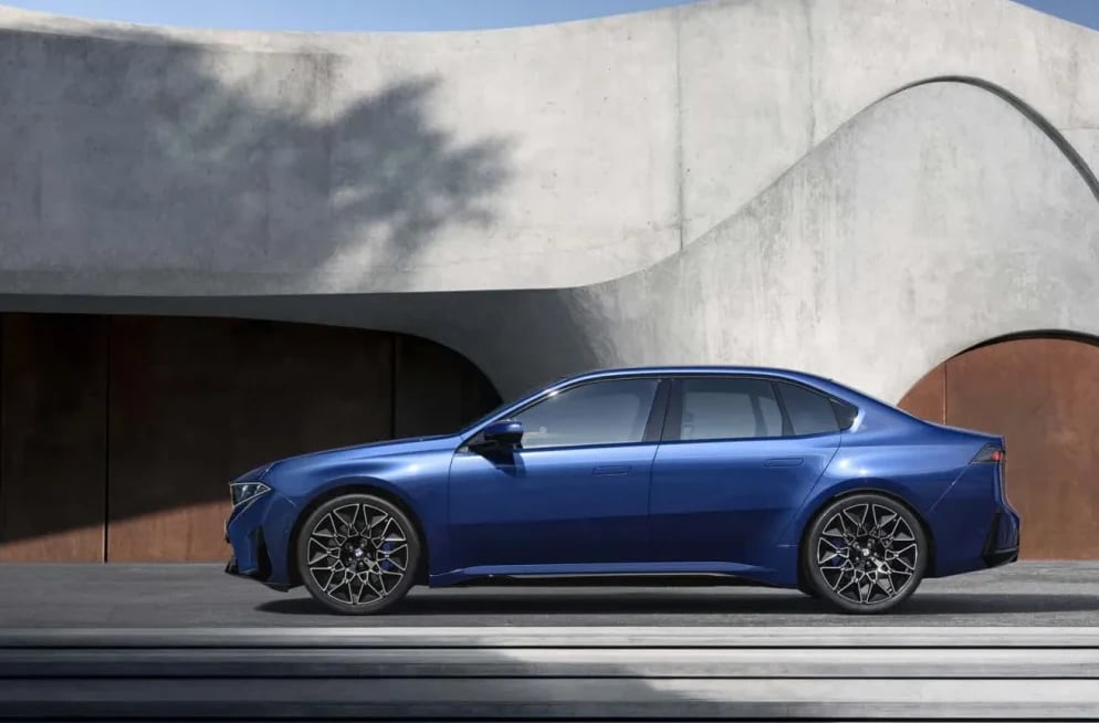 Fotografija profila plavog BMW-a i7 sa modernim dizajnom i aluminijskim felgama, pozicioniranog ispred zgrade sa zakrivljenim betonskim elementima i tamnim drvenim panelima.