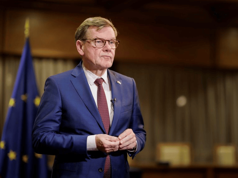 Portret starijeg muškarca u plavom odijelu i crvenoj kravati sa naočalama, sa zastavom Evropske unije u pozadini.