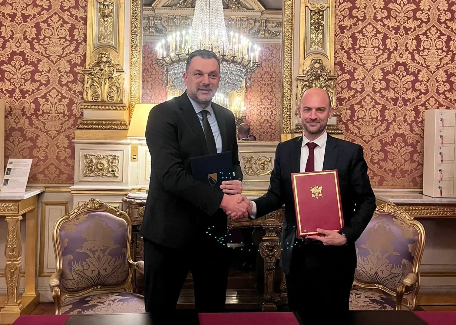 Dva muškarca u odijelima rukuju se ispred raskošnog zlatnog kamina i tapeta, držeći dokumente.