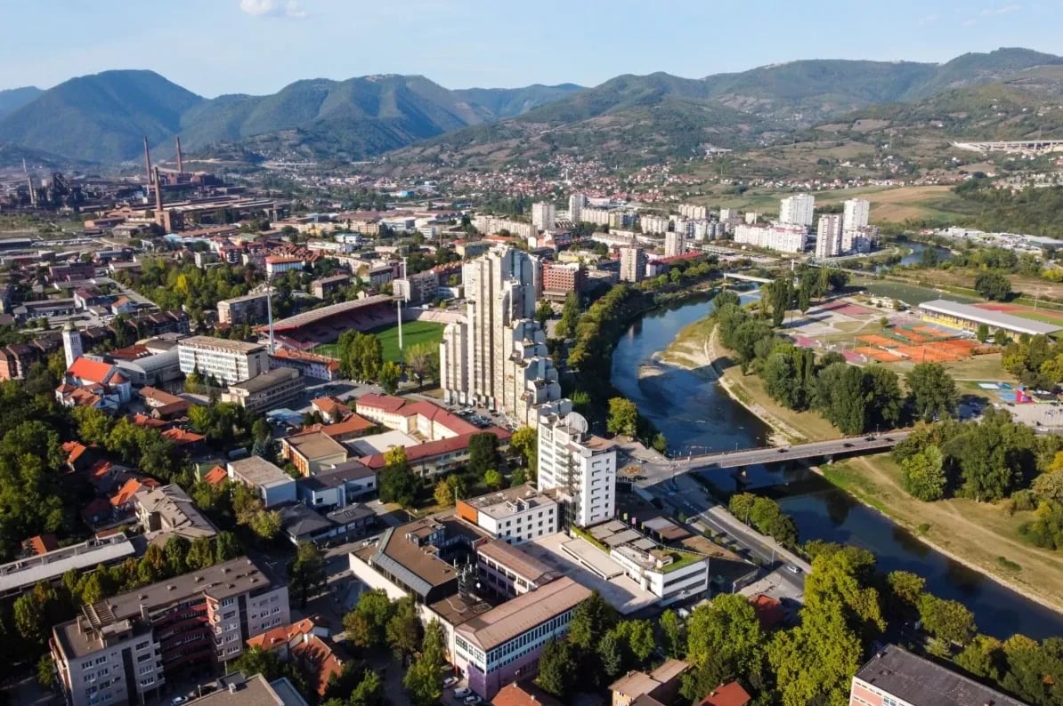 Panoramski pogled na Zenicu iz zraka, prikazuje gradsku jezgru sa rijekom Bosnom, sportskim terenima, stadionskim kompleksom i planinskim krajolikom u pozadini.