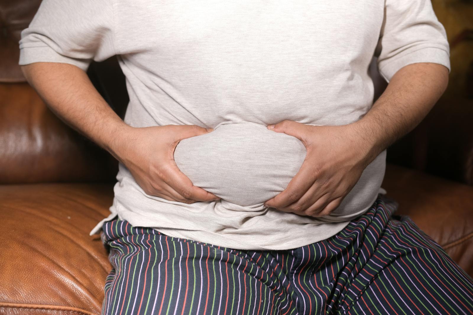 A close-up view of a man holding his tummy while sitting on a couch, highlighting lifestyle choices.