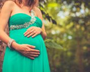 A pregnant woman wearing a green dress gently holds her belly in a serene outdoor setting.