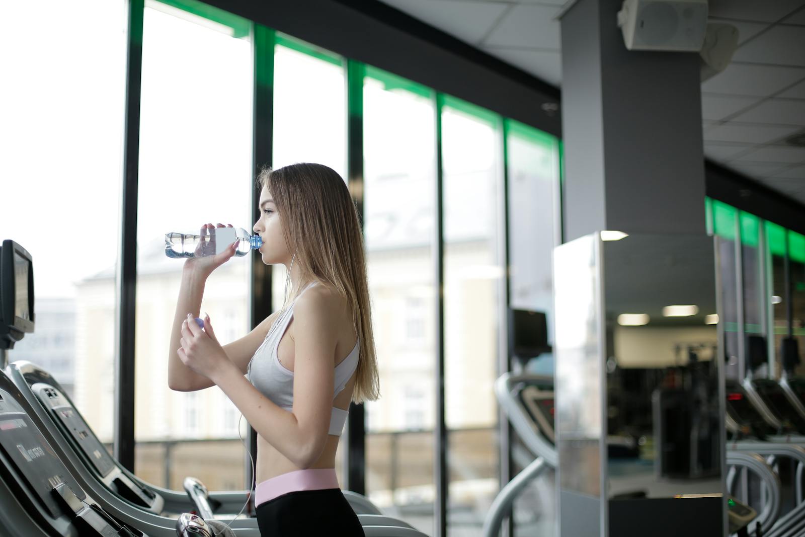 A young woman drinks water during her workout on a treadmill at a modern indoor gym.