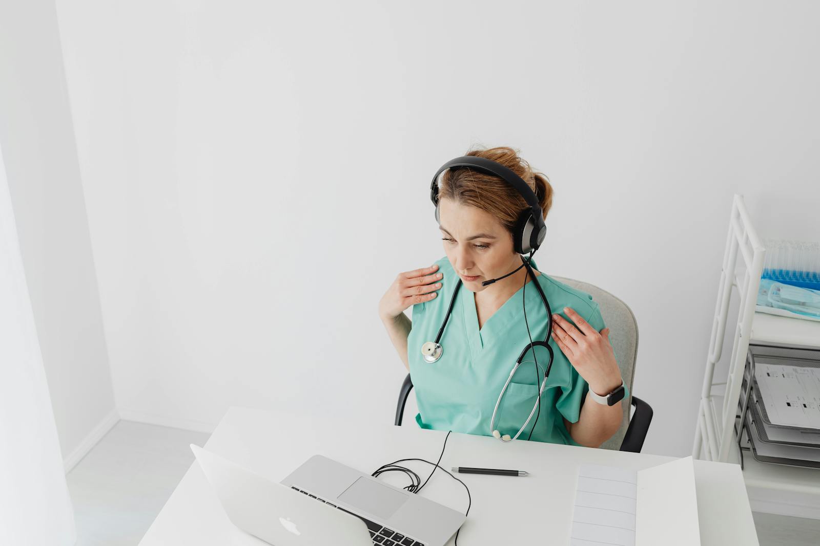 Female doctor in scrub suit conducting an online consultation using a laptop and headset in a bright office space.