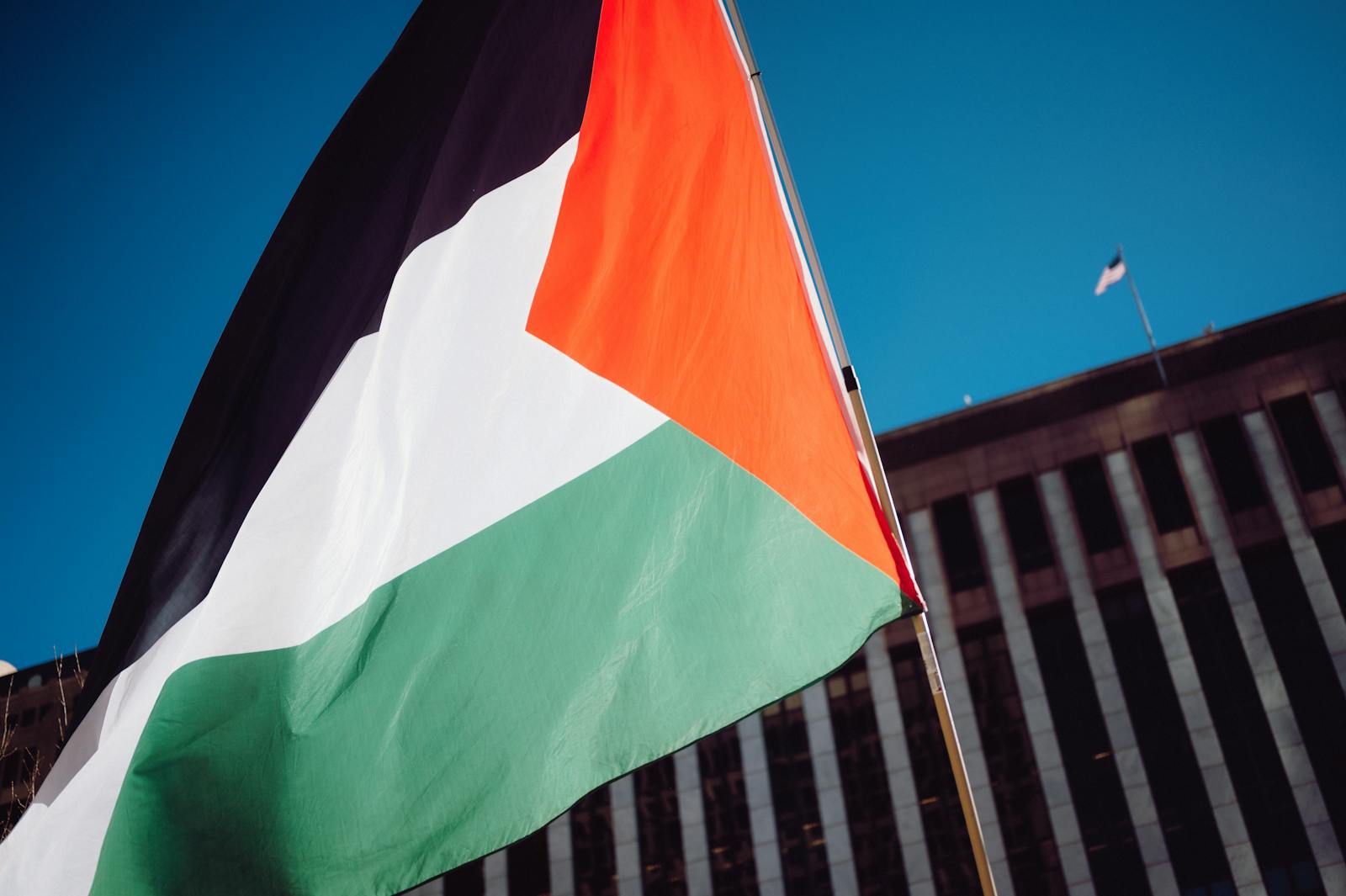 A Palestinian flag waving proudly against a blue sky with a modern building backdrop.