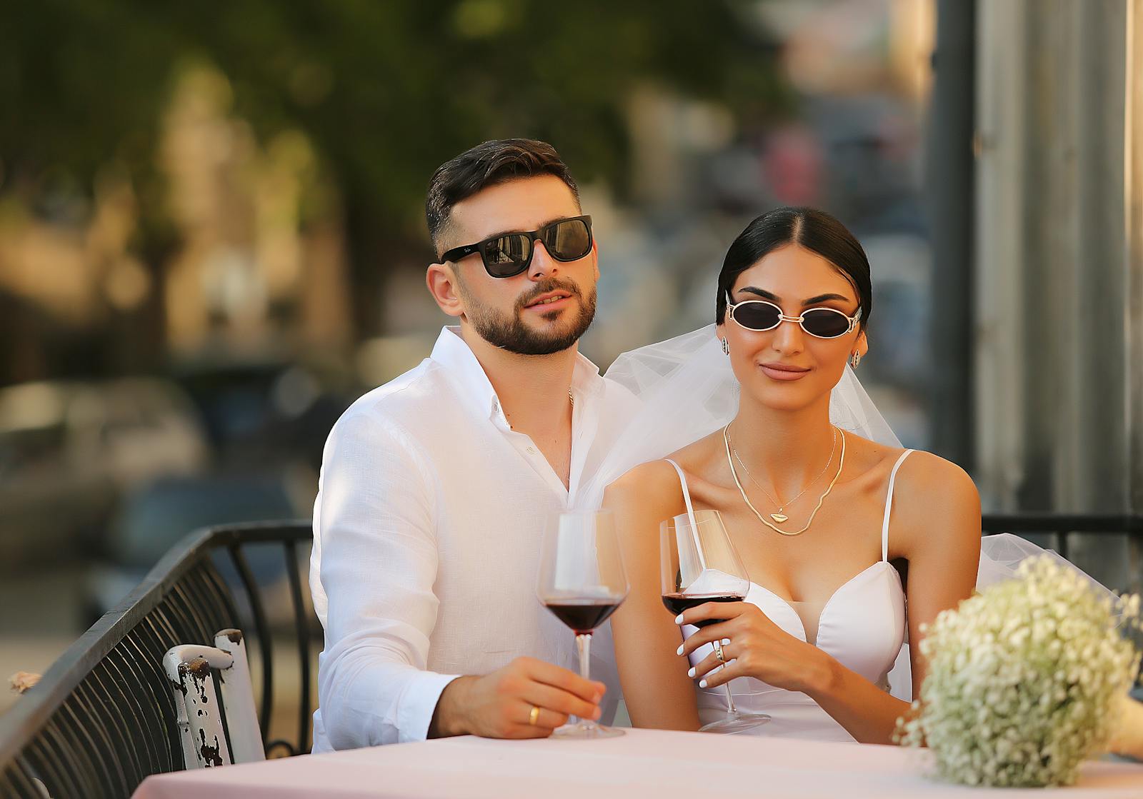 Elegant newlyweds in sunglasses toast with wine in an outdoor urban setting, exuding modern romance.
