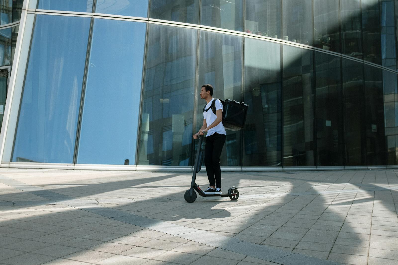 Man on electric scooter with backpack in front of modern glass building, representing urban delivery service.