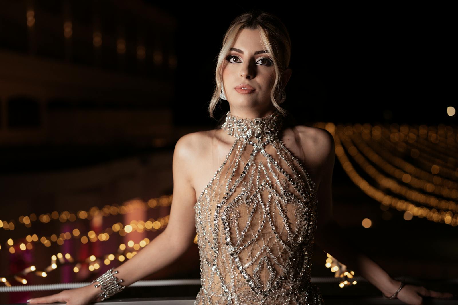 A woman in an elegant gown poses in front of twinkling lights at night, exuding sophistication.