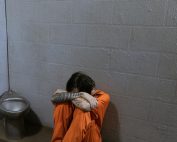 A solitary prisoner in orange uniform sits on the floor of a sparse jail cell.