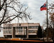A view of the Slovak Parliament building surrounded by autumn trees with the national flag waving under an overcast sky.