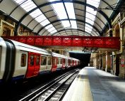 A vibrant red bridge contrasts with a sleek train at a historic railway station, showcasing travel and architecture.