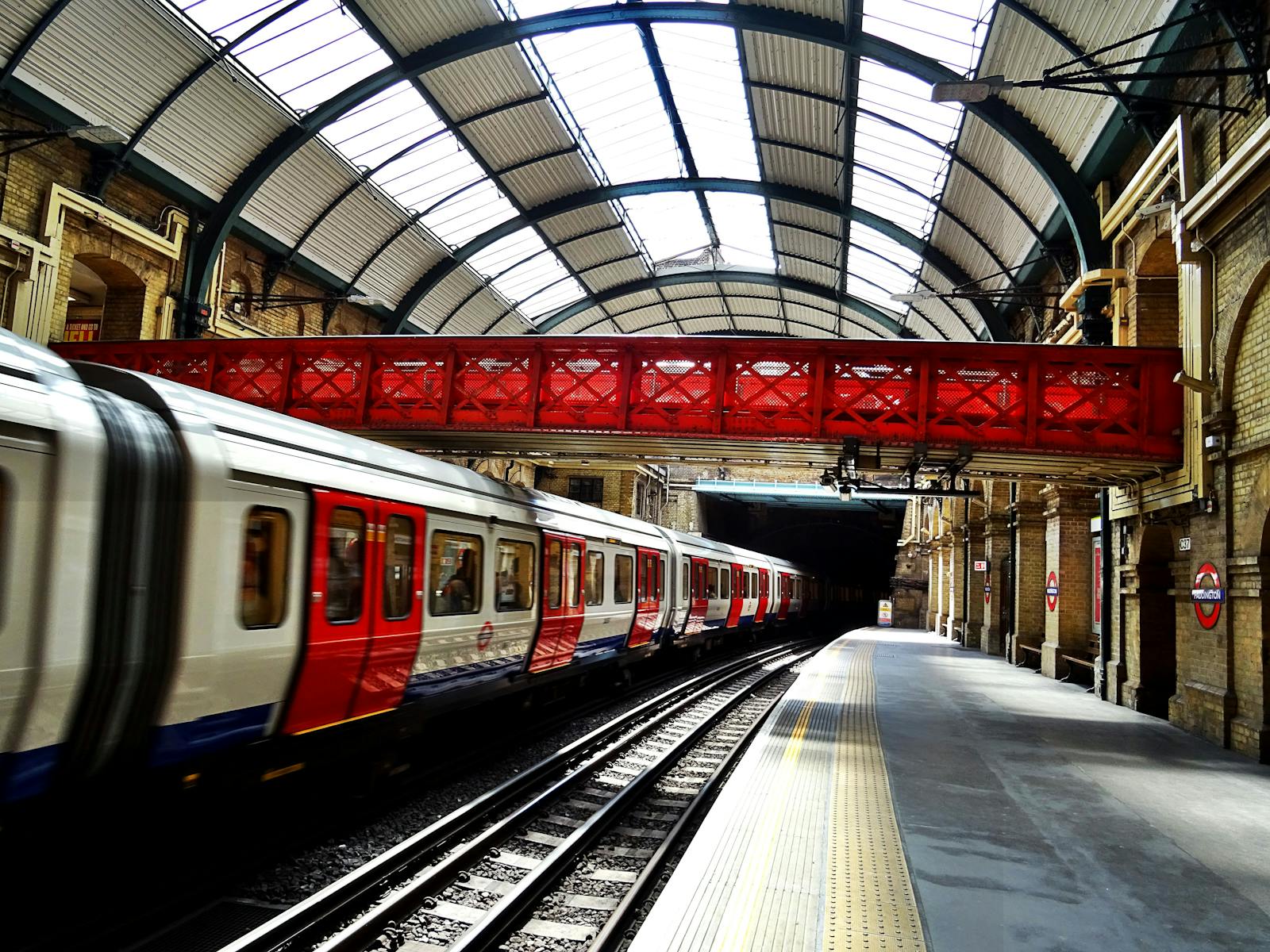 A vibrant red bridge contrasts with a sleek train at a historic railway station, showcasing travel and architecture.