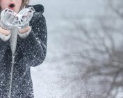 Woman in cozy winter clothing blowing snowflakes with excitement outdoors in a snowy setting.