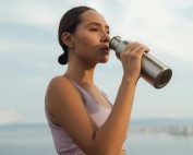A woman drinks from a metal bottle on a sunny day by the beach, wearing a sports bra.