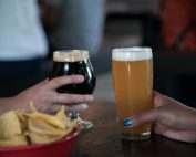 Close-up view of two hands holding beer glasses and a bowl of chips.