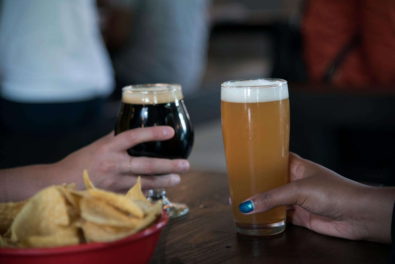 Close-up view of two hands holding beer glasses and a bowl of chips.