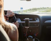 View from behind a woman driving a car on a sunny day with a phone mounted on the dashboard.