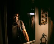 A man with a serious expression stands in front of a mirror in a dimly lit room.
