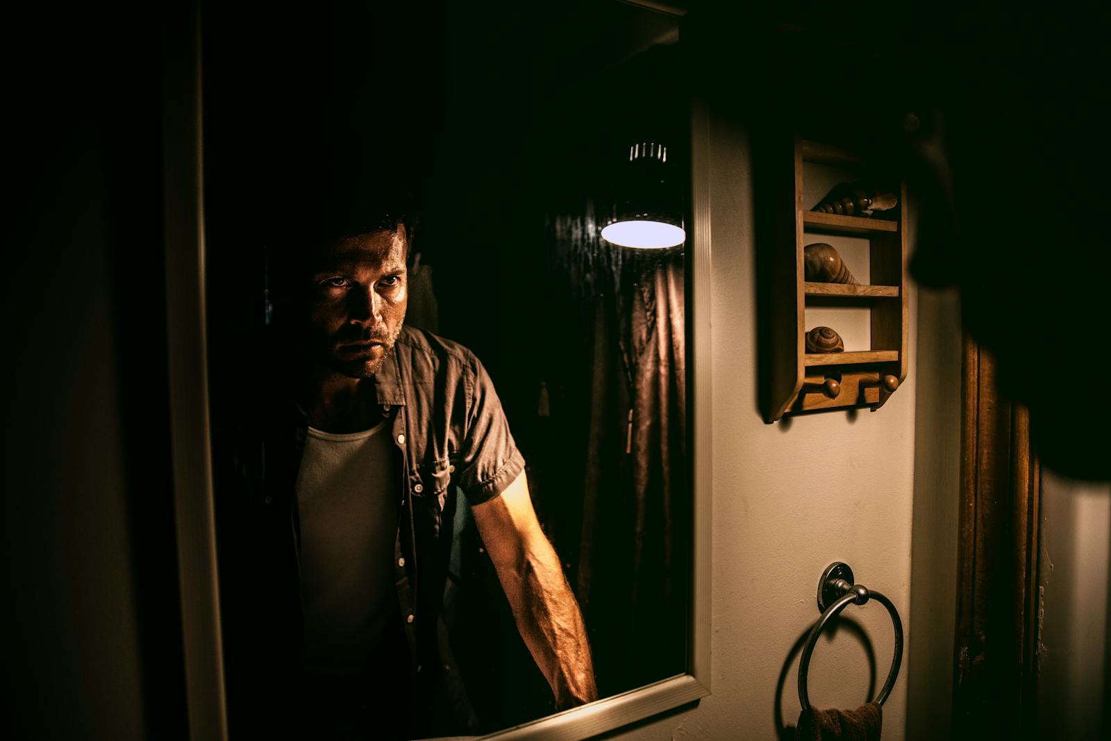 A man with a serious expression stands in front of a mirror in a dimly lit room.