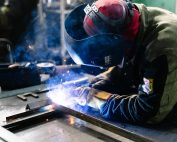 A welder works on metal fabrication indoors, creating bright sparks and smoke.