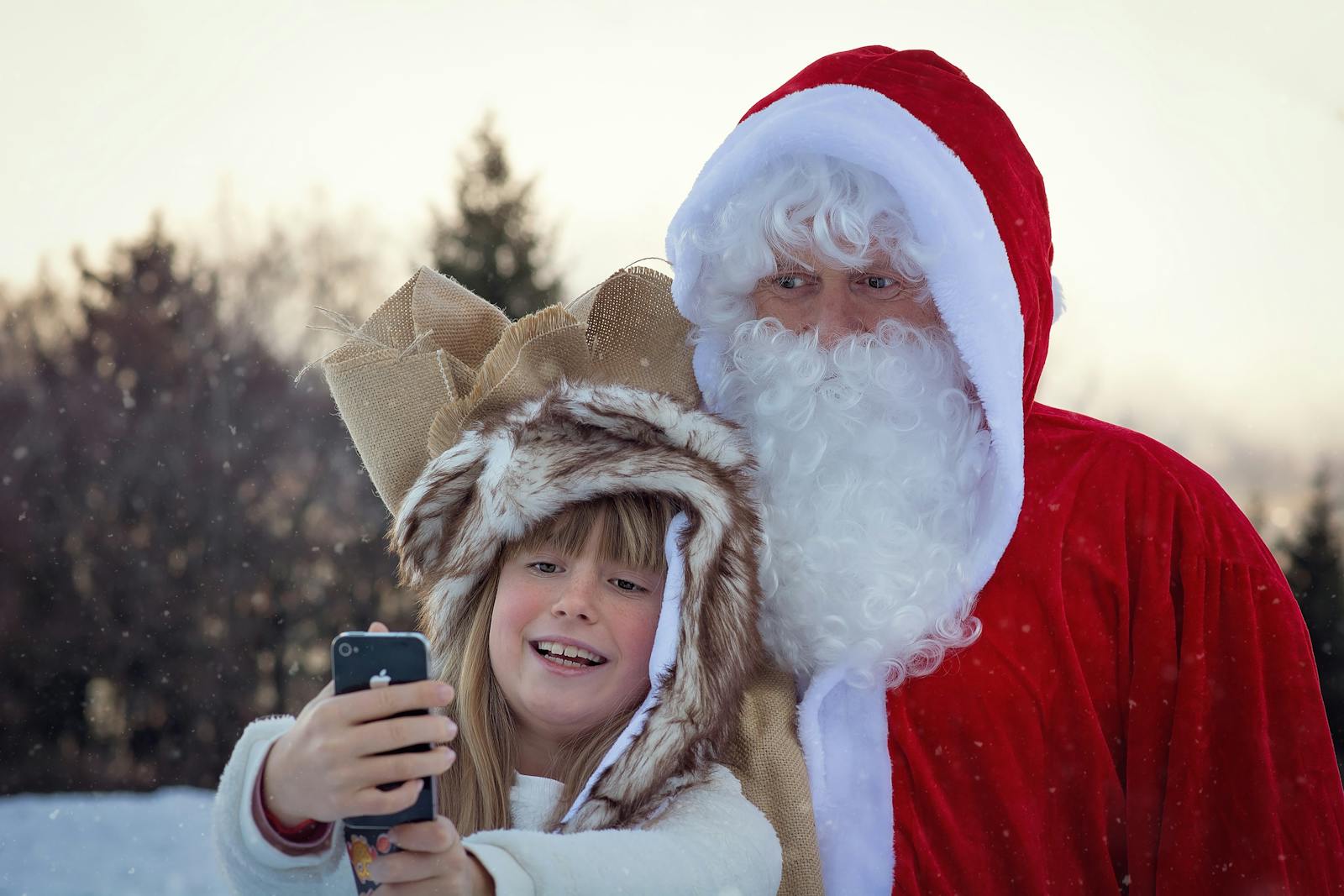 A young girl takes a selfie with Santa Claus in a snowy outdoors setting.