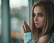 Stylish portrait of a woman with striking makeup gazing out a glass window, capturing elegance and contemplation.