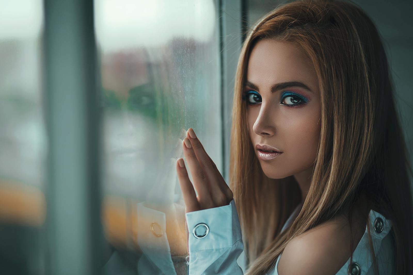 Stylish portrait of a woman with striking makeup gazing out a glass window, capturing elegance and contemplation.