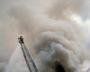 A brave firefighter combats a massive blaze from a high ladder, engulfed in thick smoke.