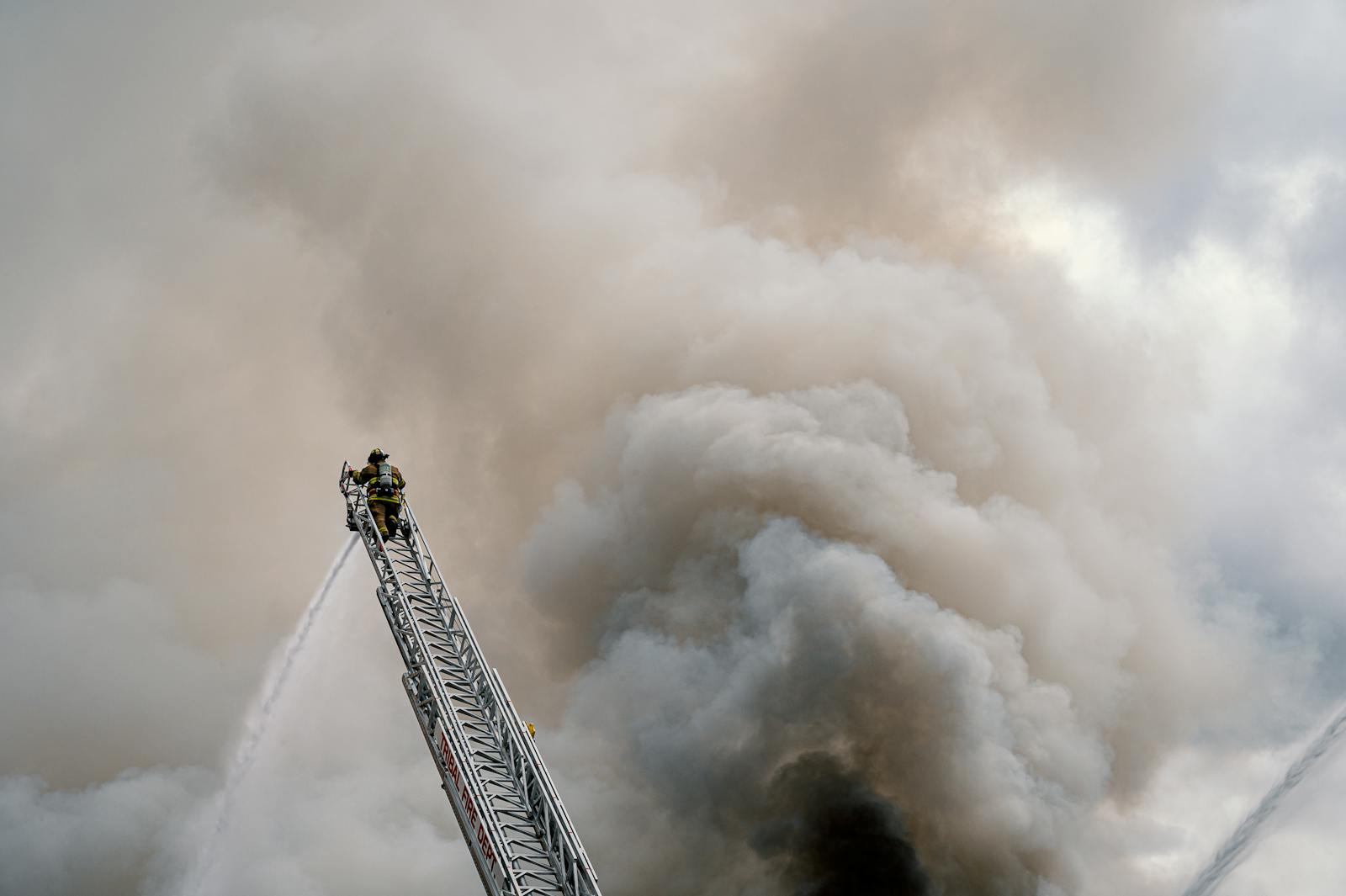 A brave firefighter combats a massive blaze from a high ladder, engulfed in thick smoke.