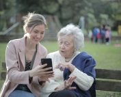 Delighted female relatives sitting together on wooden bench in park and browsing mobile phone while learning using