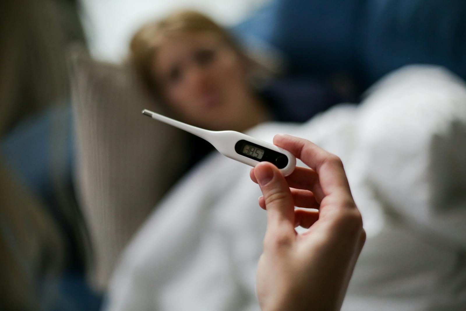 Hand holding a digital thermometer with blurred background of a sick person.