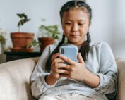 A young girl comfortably using her smartphone indoors, surrounded by plants.