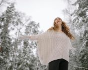 A woman enjoying a winter day in a snowy forest with outstretched arms and a joyful expression.