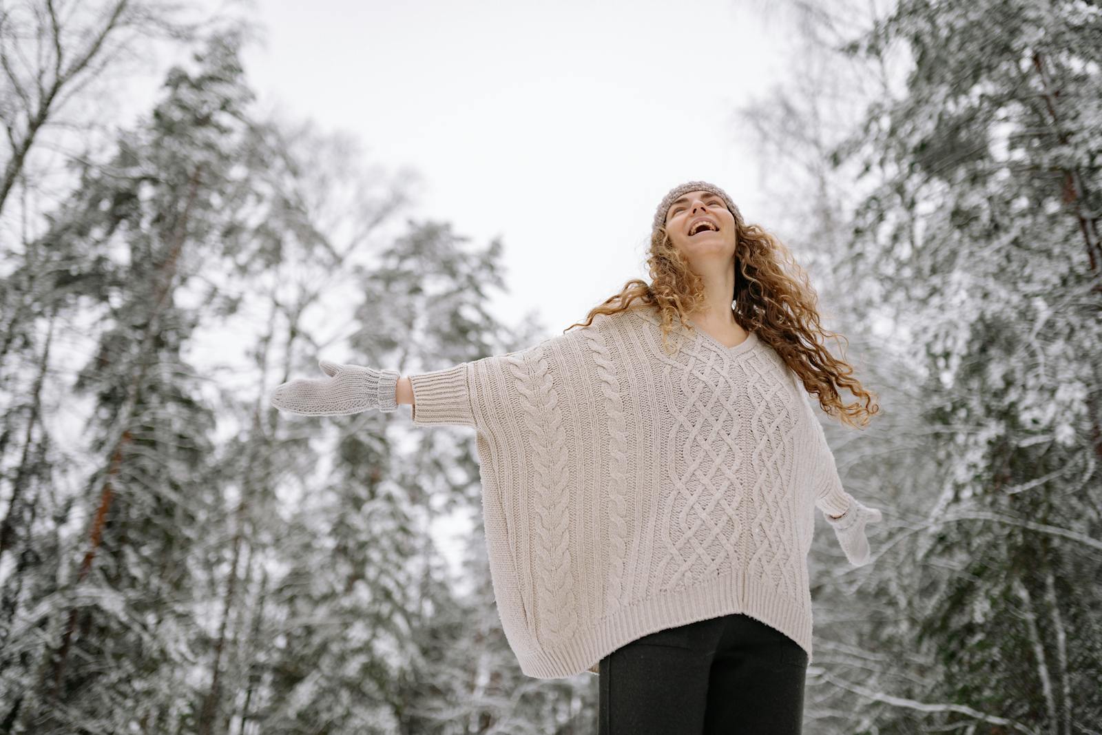 A woman enjoying a winter day in a snowy forest with outstretched arms and a joyful expression.
