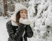 Caucasian woman blowing snow in a winter forest, wearing warm clothes.