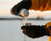 Close-up of gloved hands pouring hot drink from thermos in snowy outdoor setting.