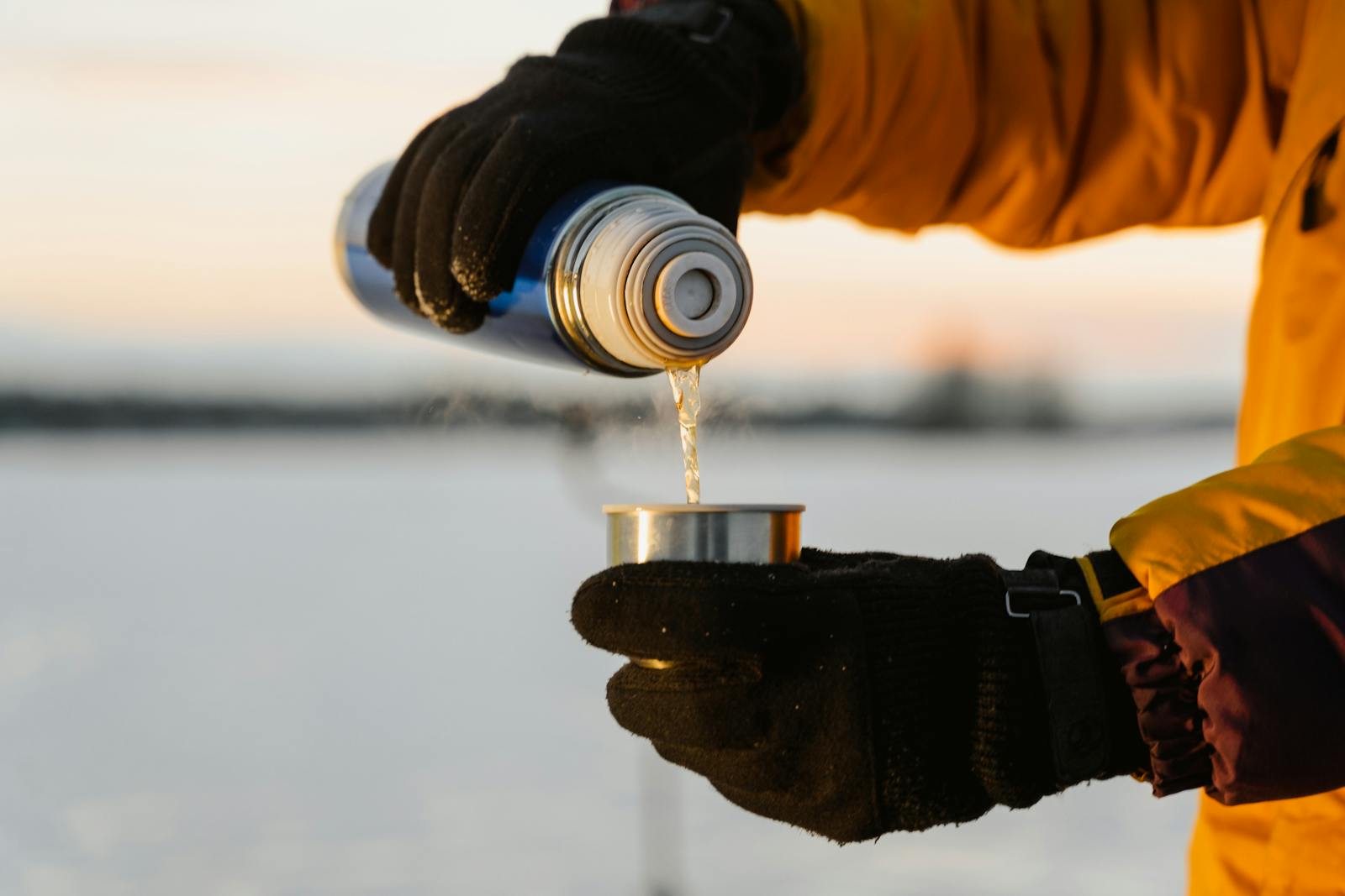 Close-up of gloved hands pouring hot drink from thermos in snowy outdoor setting.