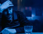 A contemplative man holds head at bar with whiskey glass, under moody lighting.