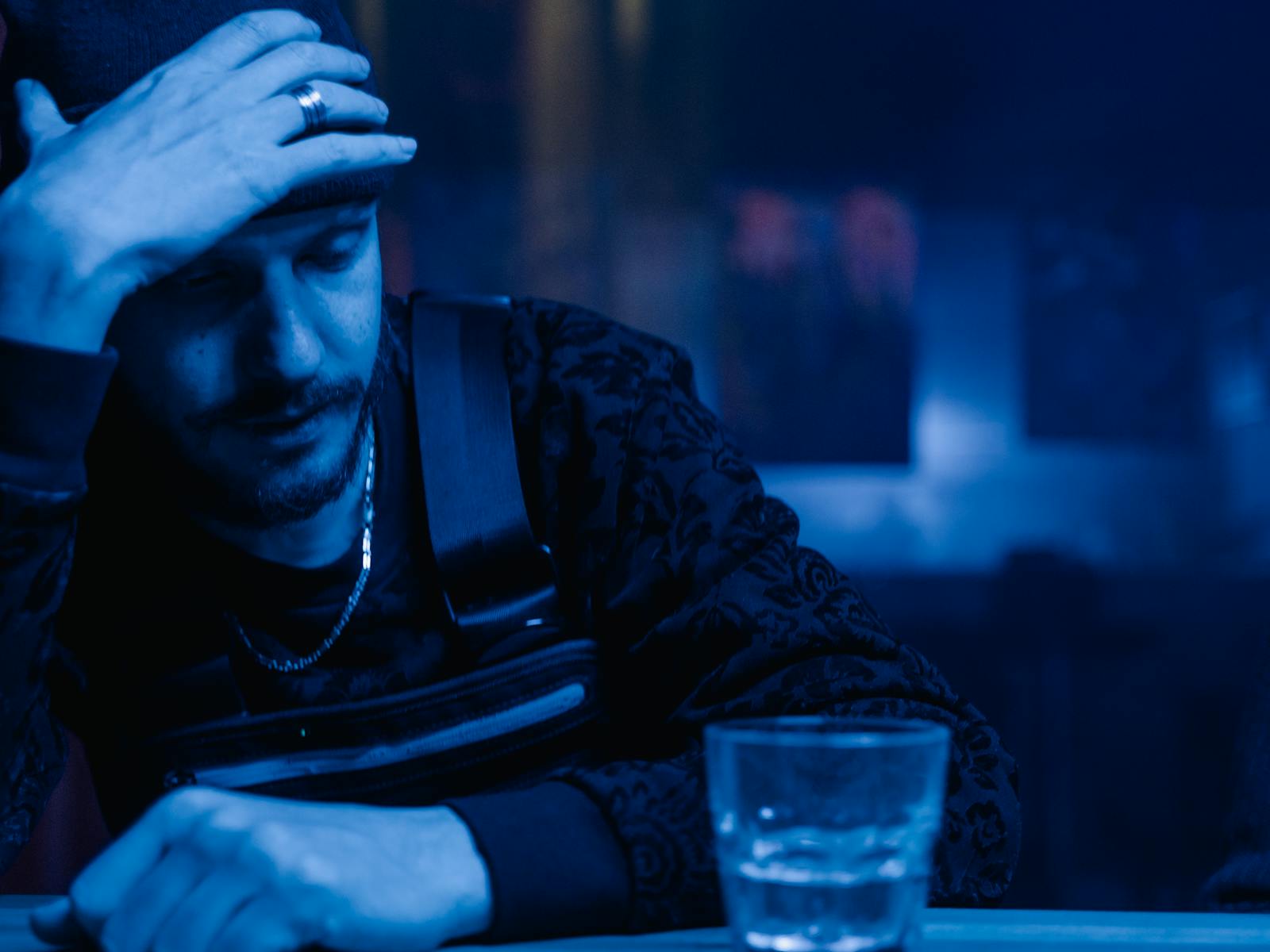 A contemplative man holds head at bar with whiskey glass, under moody lighting.
