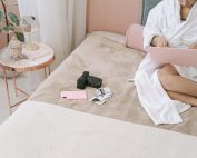 Woman in a robe using a laptop on a bed with a camera and photos nearby.