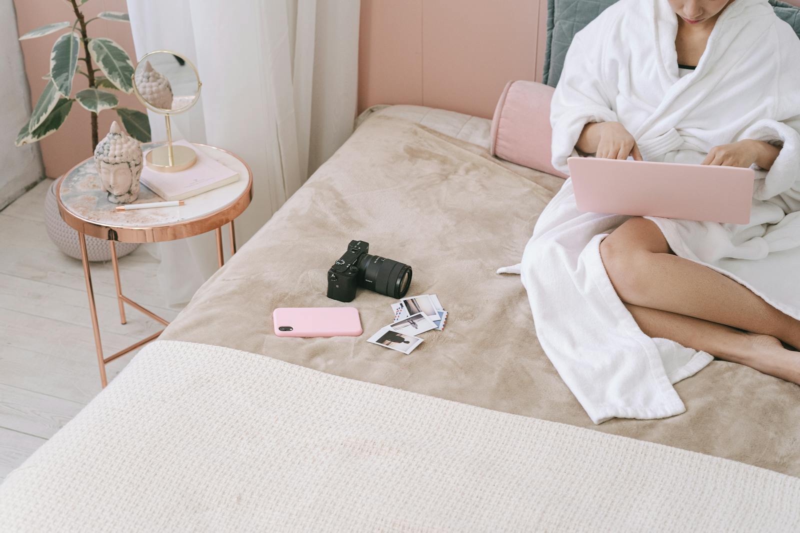 Woman in a robe using a laptop on a bed with a camera and photos nearby.