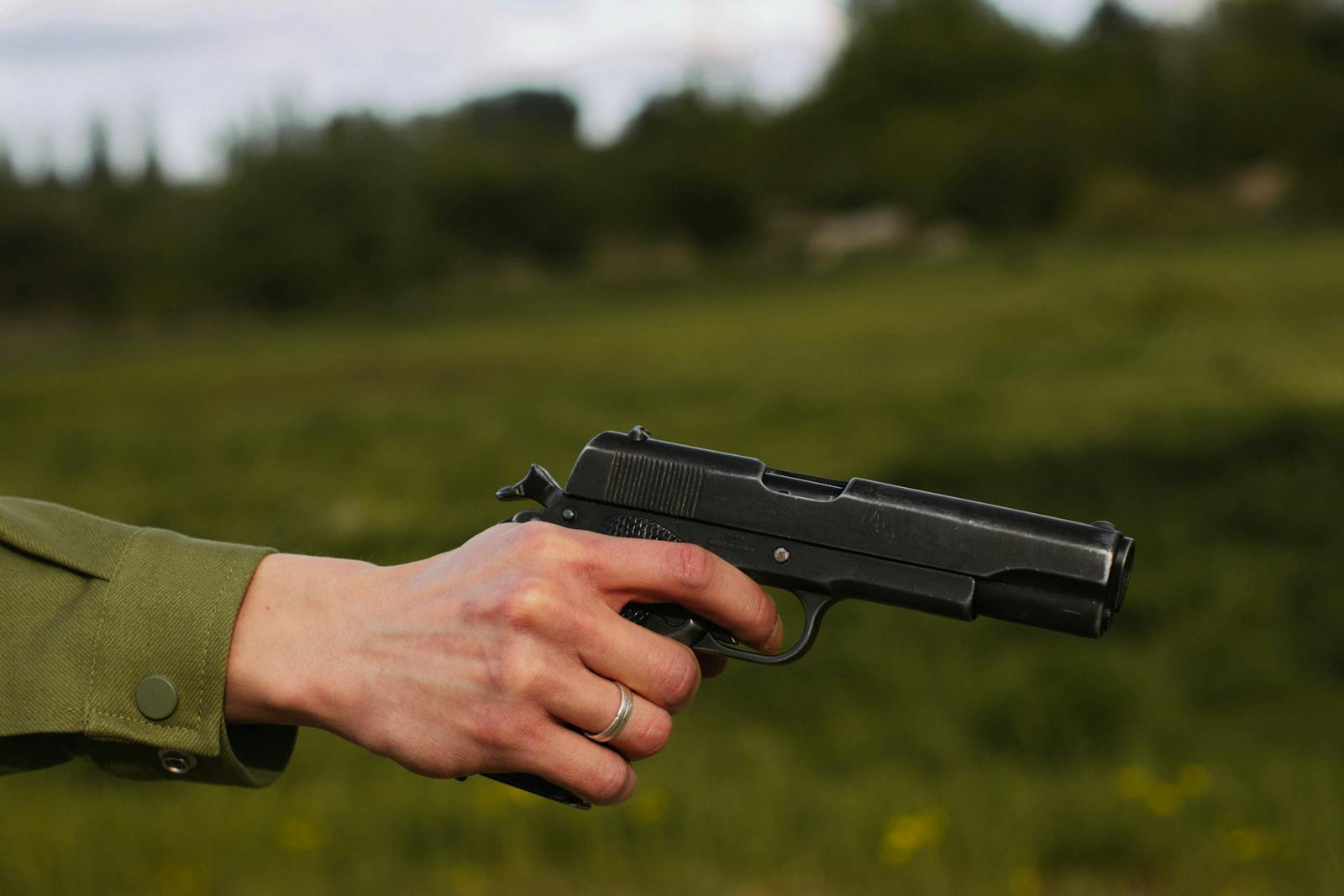 A close-up of a hand wearing a silver ring, holding a pistol outdoors.